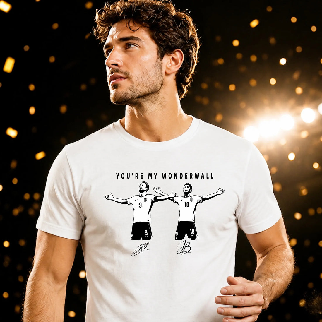 A man wearing a white graphic t-shirt with a black print of two soccer players and the text 'YOU'RE MY WONDERWALL'. The background is dark with warm golden bokeh lights.